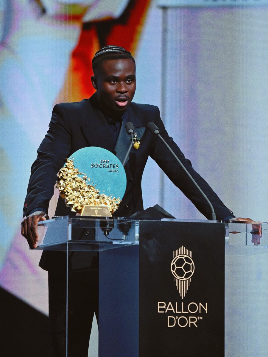 Bukayo Saka speaking at a Ballon d&rsquo;Or podium beside the 2026 Socrates Award trophy, a round teal disc with gold shards